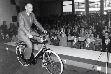 2407 De heer H. Buma rijdt op een nieuwe fiets het toneel van de aula van de G.S.G.D. op. Door de ramen is op de ...
