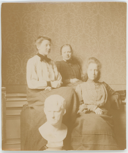 112-0015 Drie vrouwen naast een wit beeld van een mannen hoofd in een interieur, 1880-1940
