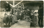 1-0007 Groepsportret van kaartspelende militairen in een barak, met een bord met de tekst De Sukkelaars Harskamp , 1920-1926